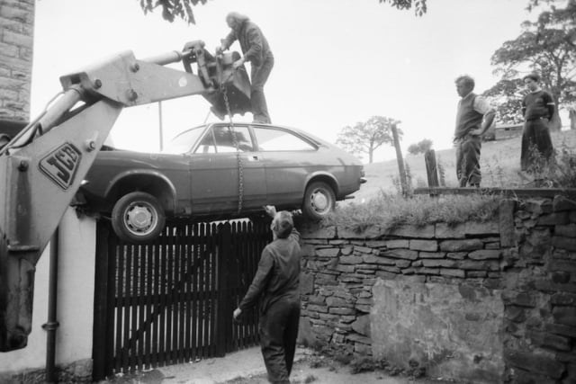 The car is chained up by Mr Alec Heap, before being lowered down from its perch on the fence.
Unwelcome guest in the village pub. A runaway car careered down a field at Cliviger and smashed into the Gordon Lennox Inn to become an unexpected gate-crasher. The Marina Saloon was left perched on a fence behind the pub and had to be rescued by a JCB with the use of chains.